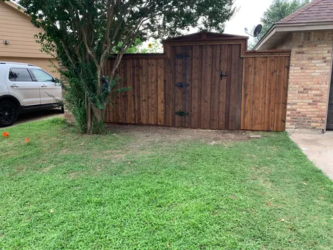 A wooden privacy fence with a decorative gate and cap, connecting a brick house to a tree line.