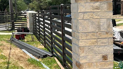 A modern horizontal metal fence under construction next to a large stone pillar.