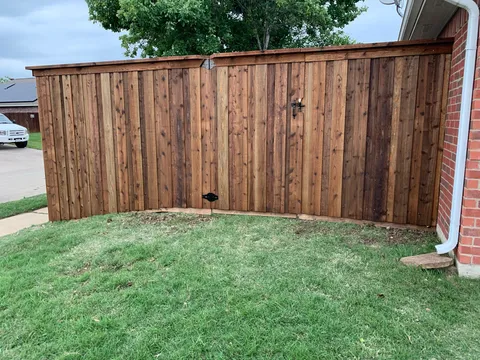 A stained wooden privacy fence with a top cap and an integrated gate, installed in a side yard next to a brick house.
