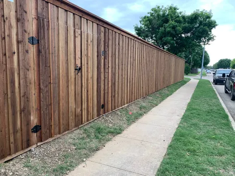 A long wooden privacy fence with vertical boards and a cap, running alongside a public sidewalk.